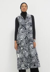 Donna che indossa un hijab nero e una maglietta nera a manica lunga sotto un abito smanicato a spirale bianco e nero, con bracciali d'argento.