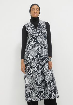 Donna che indossa un hijab nero e una maglietta nera a manica lunga sotto un abito smanicato a spirale bianco e nero, con bracciali d'argento.