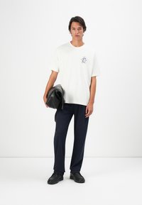 Junger Mann in einem weißen T-Shirt mit Grafik und dunklen Hosen, der einen schwarzen Leder-Rucksack hält, steht vor einem schlichten weißen Hintergrund.