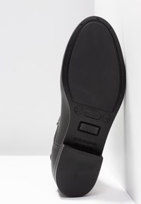 Chaussure en cuir noir avec une semelle en caoutchouc texturée. Présente un logo en relief, des marquages de taille et un design supérieur lisse. Fabriquée en Espagne.