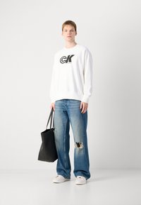 Felpa bianca con colletto a girocollo e logo nero, jeans blu strappati e sneakers bianche. Porta una borsa tote nera.