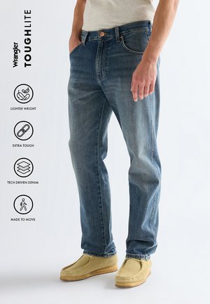 Mann trägt hellblaue Wrangler-Jeans und beige Mokassinschuhe, steht mit teils in den Taschen steckenden Händen vor einfarbigem Hintergrund.