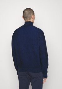 Dunkelblauer Strickpullover mit vertikal gerippter Struktur, hohem Hals und enganliegenden Bündchen. Der Stoff wirkt weich und strukturiert.