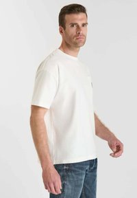 Uomo con capelli corti castani, indossa una semplice camicia bianca a maniche corte e jeans blu, in piedi davanti a uno sfondo grigio chiaro.