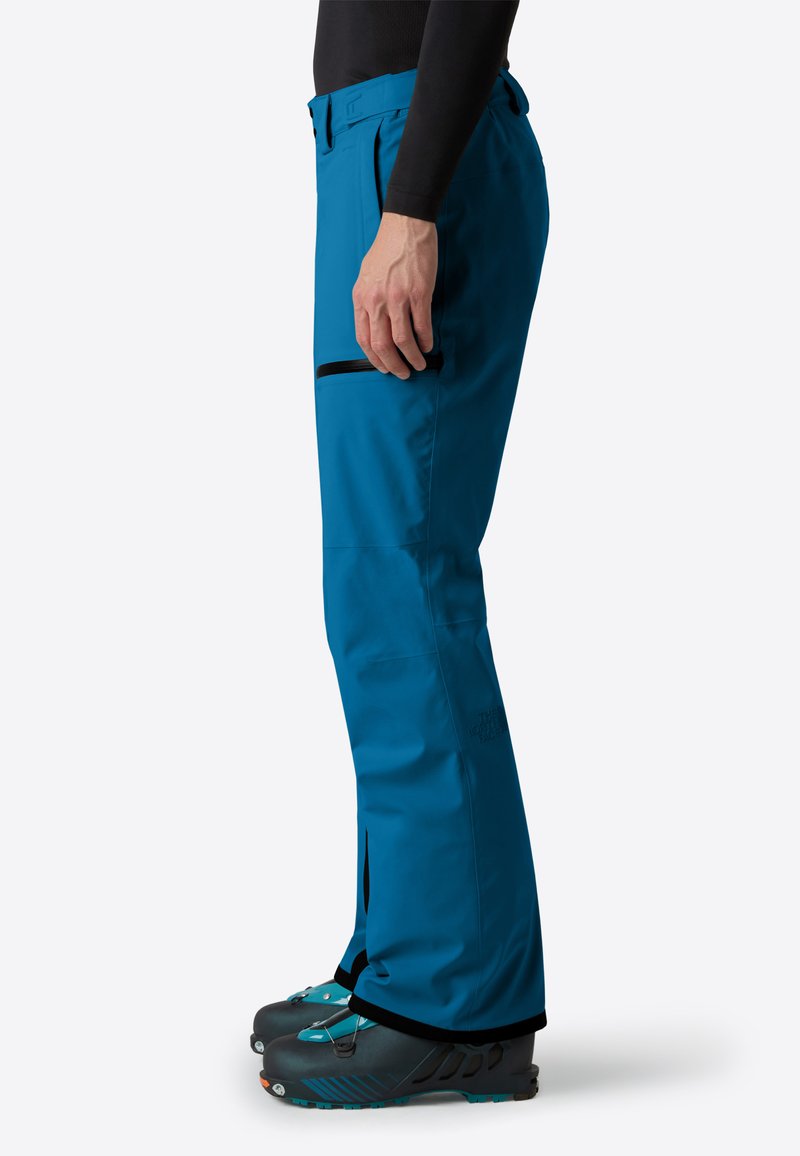 Persona che indossa pantaloni da esterno di un brillante blu con una tasca con zip sulla coscia e scarpe da trekking nere e blu.