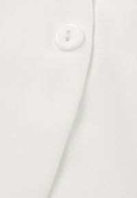 Gros plan sur un tissu blanc montrant un bouton blanc à deux trous cousu avec un chevauchement de tissu, probablement une partie d'une fermeture de vêtement.