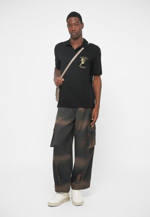 Młody mężczyzna w czarnej koszulce polo z złotym nadrukiem, w czarno-brązowych spodniacach typu cargo w tie-dye, białych sneakersach i be�żowej torbie na ramię stoi.