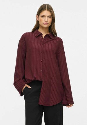 Jeune femme portant une chemise ample à manches longues rayée bordeaux et un pantalon noir, une main dans la poche, sur un fond uni.