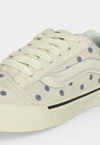 Λευκός-μπεζ αθλητικός με κρεμ κορδόνια, μικρό μπλε και πορτοκαλί λουλουδάτο σχέδιο, λευκή κυματιστή ρίγα και χοντρή λαστιχένια σόλα με μαύρη γραμμή.