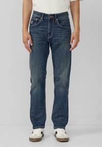 Niebieskie dżinsy mają prosty krój nogawki, lekkie przetarcia, miedziane szwy i zapięcie na guziki. Noszone z białymi i brązowymi trampkami.