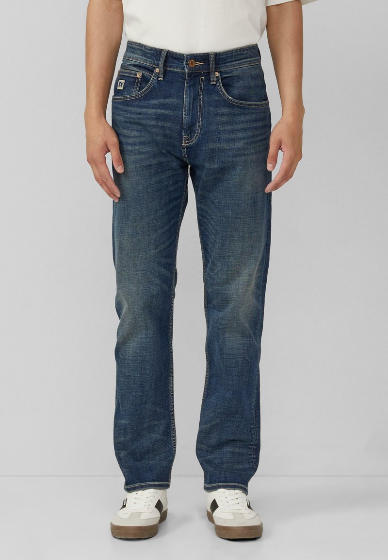 Niebieskie dżinsy mają prosty krój nogawki, lekkie przetarcia, miedziane szwy i zapięcie na guziki. Noszone z białymi i brązowymi trampkami.