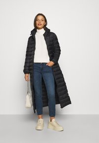 Doudoune noire avec un design long et matelassé, dotée d'une capuche, de zips latéraux et d'une ouverture devant. Portée par-dessus un pull blanc et un jean bleu.