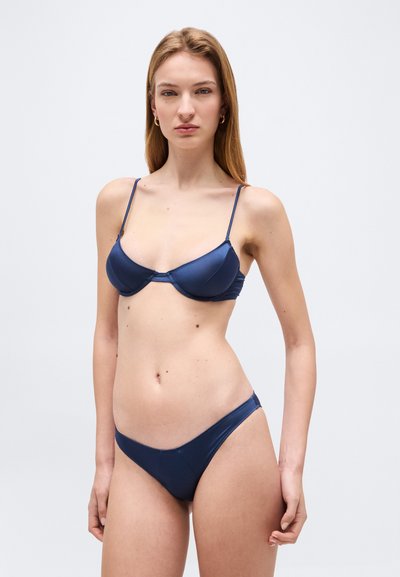 Νεαρή γυναίκα με μακριά ξανθά μαλλιά που φοράει μπλε σκούρο μπικίνι με λεπτές τιράντες μπροστά σε απλό ανοιχτό φόντο.