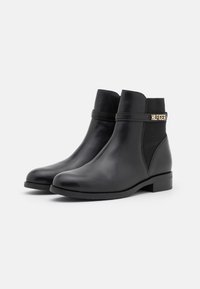 Une paire de bottines noires en cuir avec des talons bas, des panneaux latéraux élastiques et une sangle décorative arborant un logo en métal "HILFIGER".