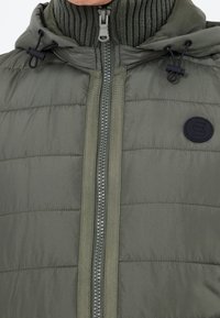 Gilet rembourré olive avec un col montant, fermeture éclair complète et accents en tricot. Il présente un badge logo noir et une capuche ajustable avec cordon.