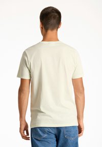Camiseta de algodón verde claro con mangas cortas, cuello redondo y una etiqueta en el lado inferior, combinada con jeans de mezclilla azules.