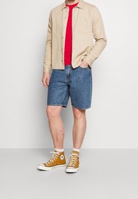 Béžová košile s dlouhým rukávem přes červené tričko, modré džínové kraťasy a žluté tenisky s vysokým svrškem, bílou podrážkou a logem.