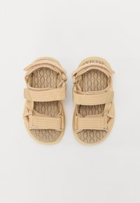 Beige Sandalen mit strukturierten Gummisohlen, verstellbaren Stoffriemen und einem runden, geschlossenen Zehenbereich. Die Riemen haben eine gewebte Textur.
