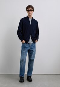 Granatowa kurtka na zamek, szara koszulka, jasnoniebieskie d�żinsy i czarne buty. Model nosi ciemne okulary przeciwsłoneczne, stoi na tle jednolitego tła.