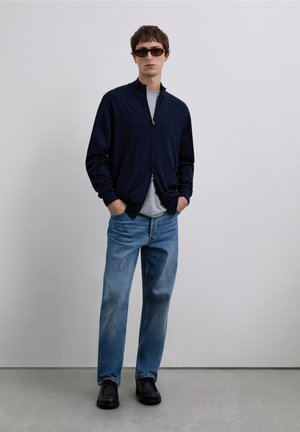 Chaqueta azul marino con cremallera, camiseta gris, vaqueros azul claro y zapatos negros. Modelo con gafas de sol oscuras, posando frente a un fondo liso.