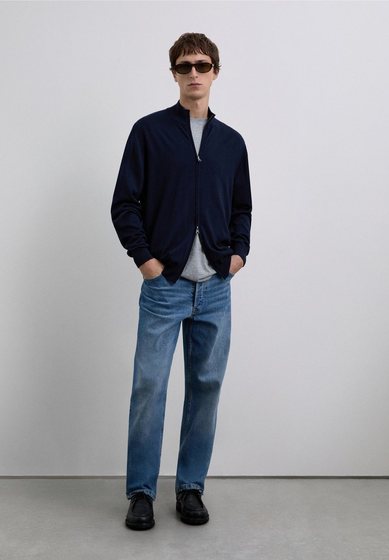 Granatowa kurtka na zamek, szara koszulka, jasnoniebieskie dżinsy i czarne buty. Model nosi ciemne okulary przeciwsłoneczne, stoi na tle jednolitego tła.