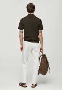 Hnědá polokošile s krátkým rukávem, bílé slim-fit kalhoty, hnědý kožený batoh a černé sandály. Viditelné tetování na pažích.