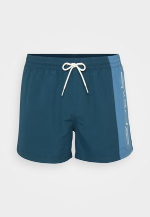 Męskie szorty kąpielowe w ciemnym odcieniu teal z jasnoniebieskim bocznym panelem. Posiadają elastyczny pas z białym sznurkiem i nadrukowane logo po boku. Gładka tekstura.