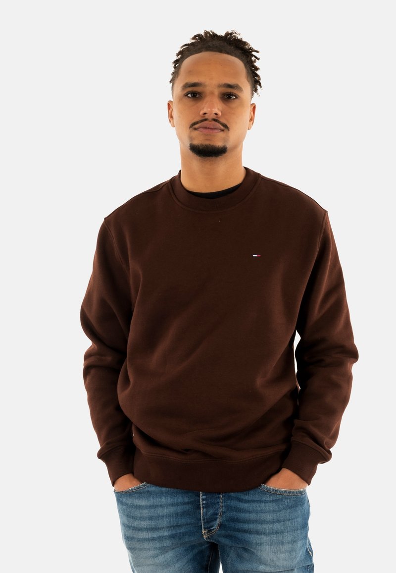 Bruine sweatshirt met een ronde hals en geribbelde manchetten, voorzien van een klein vlaggenlogo op de borst en gecombineerd met blauwe spijkerbroeken.