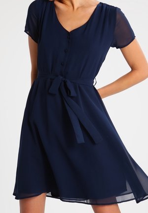 Femme portant une robe bleu marine à manches courtes, longueur au genou, avec un col en V, des boutons à l'avant et une ceinture nouée à la taille.