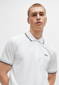 Tricou polo alb din țesătură texturată, având o deschidere cu trei butoane, logo brodat negru și guler și mâneci cu margine neagră.