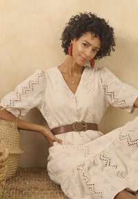Abito bianco con ricami a zigzag e bottoni, stretto in vita da una cintura marrone con una fibbia decorativa. Cesto intrecciato nelle vicinanze.