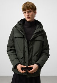 Zielono-oliwkowa kurtka ocieplana z zamkiem błyskawicznym i zapięciem na napy, regulowany kaptur, dwie przednie kieszenie, noszona na czarnym golfie. Ręce trzymają okulary przeciwsłoneczne.