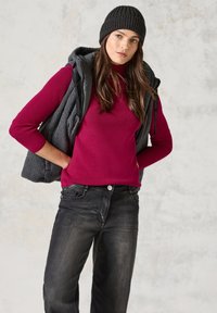 Bordeauxroter gerippter Pullover unter einer grauen Fleeceweste, getragen mit schwarzen Jeans. Das Model trägt eine gestrickte schwarze Mütze und hat eine lässige Pose. Der Hintergrund ist hell.