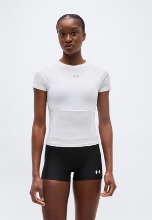 Femme debout portant un maillot de sport blanc ajusté et un short noir avec des logos Under Armour sur un fond uni.