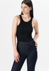 Νέα γυναίκα με ίσια καστανά μαλλιά, φορώντας ένα αμάνικο μαύρο τοπ και σκούρα μπλε ψηλόμεσα τζιν, ποζάροντας μπροστά σε λευκό φόντο.