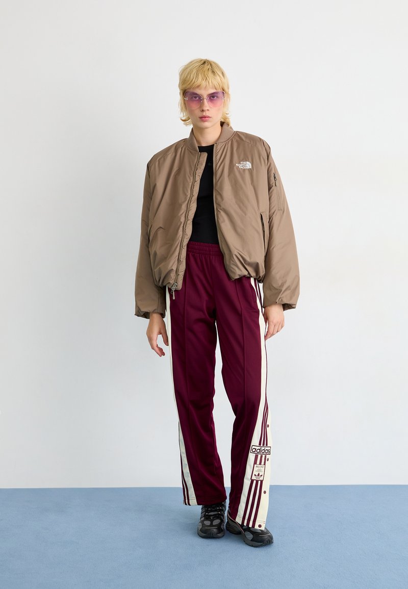 Giacca bomber isolata marrone con una maglietta nera, abbinata a pantaloni tuta borgogna con strisce bianche e logo Adidas. Calzature nere.