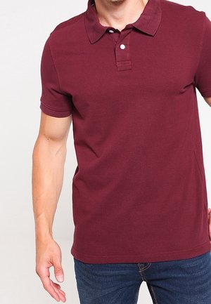 Homme portant un polo manches courtes bordeaux avec deux boutons, associé à un jean bleu, debout devant un fond uni.