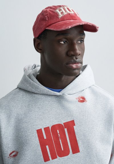 Rød caps med hvite bokstaver "HOT", grå hettegenser med fet rød "HOT"-trykk, med et leppestiftavtrykk på skulderen.