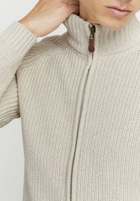 Suéter de punto beige con cierre, cuello alto, patrón texturizado y un tirador de cremallera de cuero marrón.