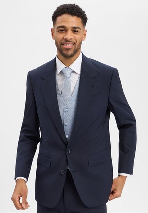 Man wearing navy suit with light blue patterned waistcoat and matching tie, smiling against plain light background.