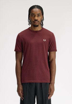 Uomo con trecce che indossa una maglietta color borgogna semplice con un piccolo logo bianco sul petto e pantaloni neri su sfondo bianco.