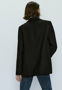 Blazer noir avec une coupe ajustée, des épaules structurées et une fente au dos. Le matériau semble lisse, associé à un jean bleu foncé.
