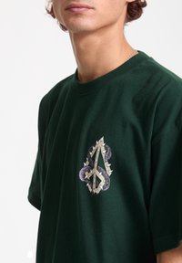 Mørkegrøn bomulds t-shirt med et grafisk motiv af sammenflettede tentakler og pigge i lilla, sølv og hvid på venstre bryst.