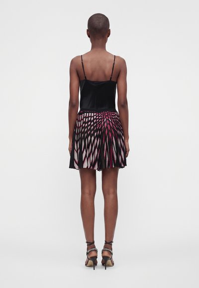 Personne aux cheveux courts portant un haut noir sans manches, une jupe plissée à motifs rose et noir, et des sandales noires à talons hauts avec des brides à la cheville.