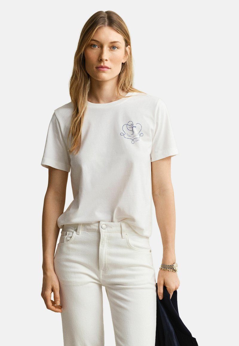 Femme aux longs cheveux blonds portant un t-shirt blanc avec un motif brodé bleu, un pantalon blanc, tenant une veste sombre dans une main.