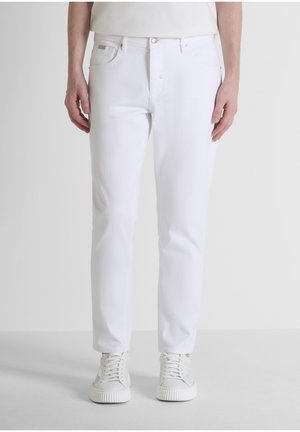 Persona che indossa jeans bianchi a taglio slim e sneaker bianche con lacci, in piedi davanti a uno sfondo grigio chiaro uniforme.