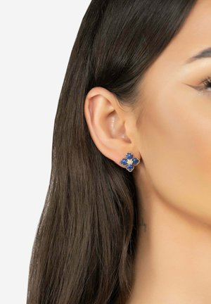 Boucles d'oreilles florales dorées ornées de pierres bleues et de perles blanches, intégrées dans un motif décoratif, reposant sur l'oreille d'un modèle.