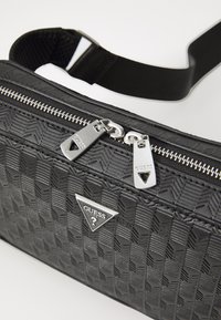 Black textured handbag with a woven pattern, silver zip closure, and branded triangular logo. Adjustable black strap for carrying.