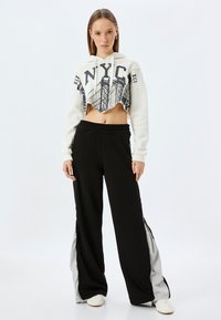 Gekürzter weißer Hoodie mit schwarzem NYC-Graphic; kombiniert mit schwarzen weiten Hosen mit grauen Seitenteilen und Reißverschlüssen. Weiße Sneakers.