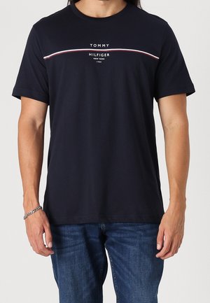 Mann in einem schwarzen Tommy Hilfiger T-Shirt mit einem weißen und roten Streifen über der Brust und blauen Jeans, der vor einem einfarbigen Hintergrund steht.